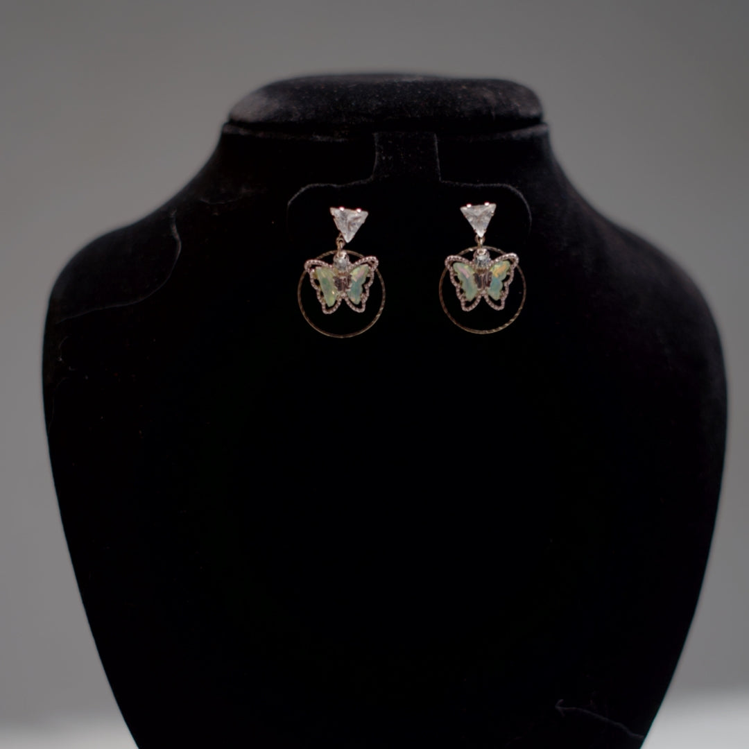 A pair of butterfly hoop drop earrings featuring green enamel and rose gold trim, displayed on a black velvet stand.
