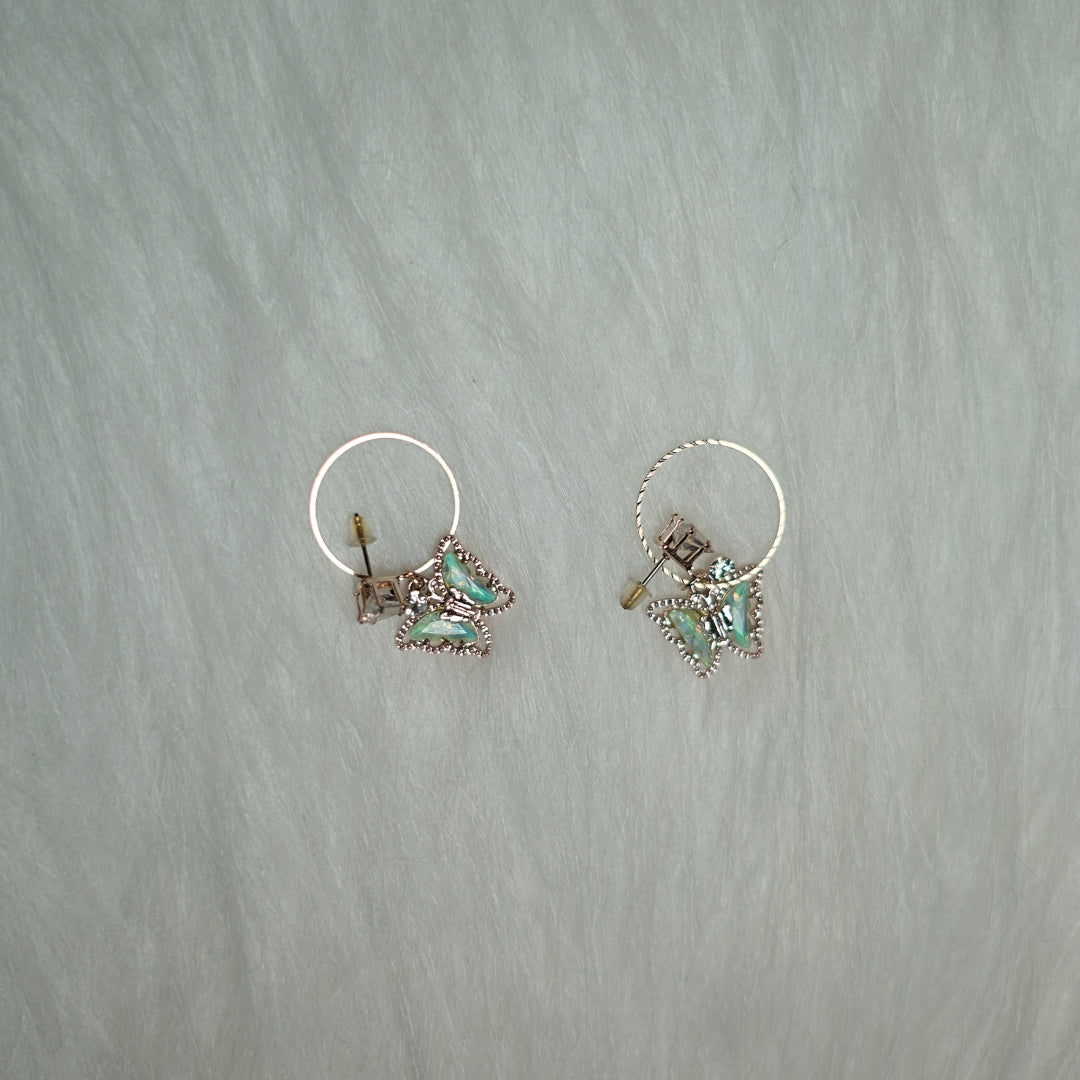 Rose gold hoop earrings with iridescent butterfly charms laid on a soft fabric background.