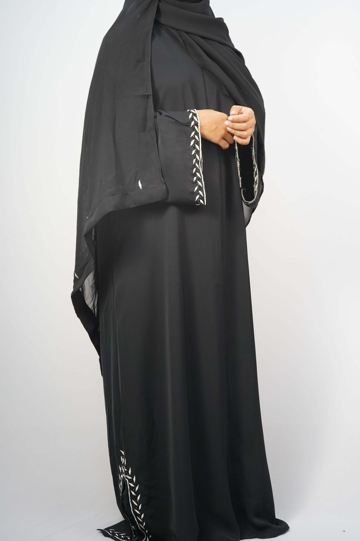 A model wearing the black abaya, highlighting the elegant fit and the contrasting white embroidery on the sleeves and hem.