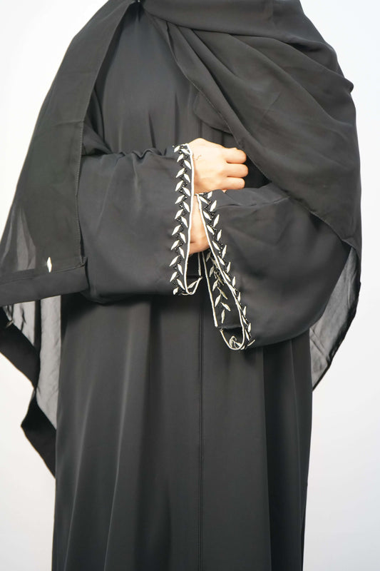 A woman wearing the Korean Nidha Abaya in black, showing the elegant fit and the detailed white embroidery on the sleeves.
