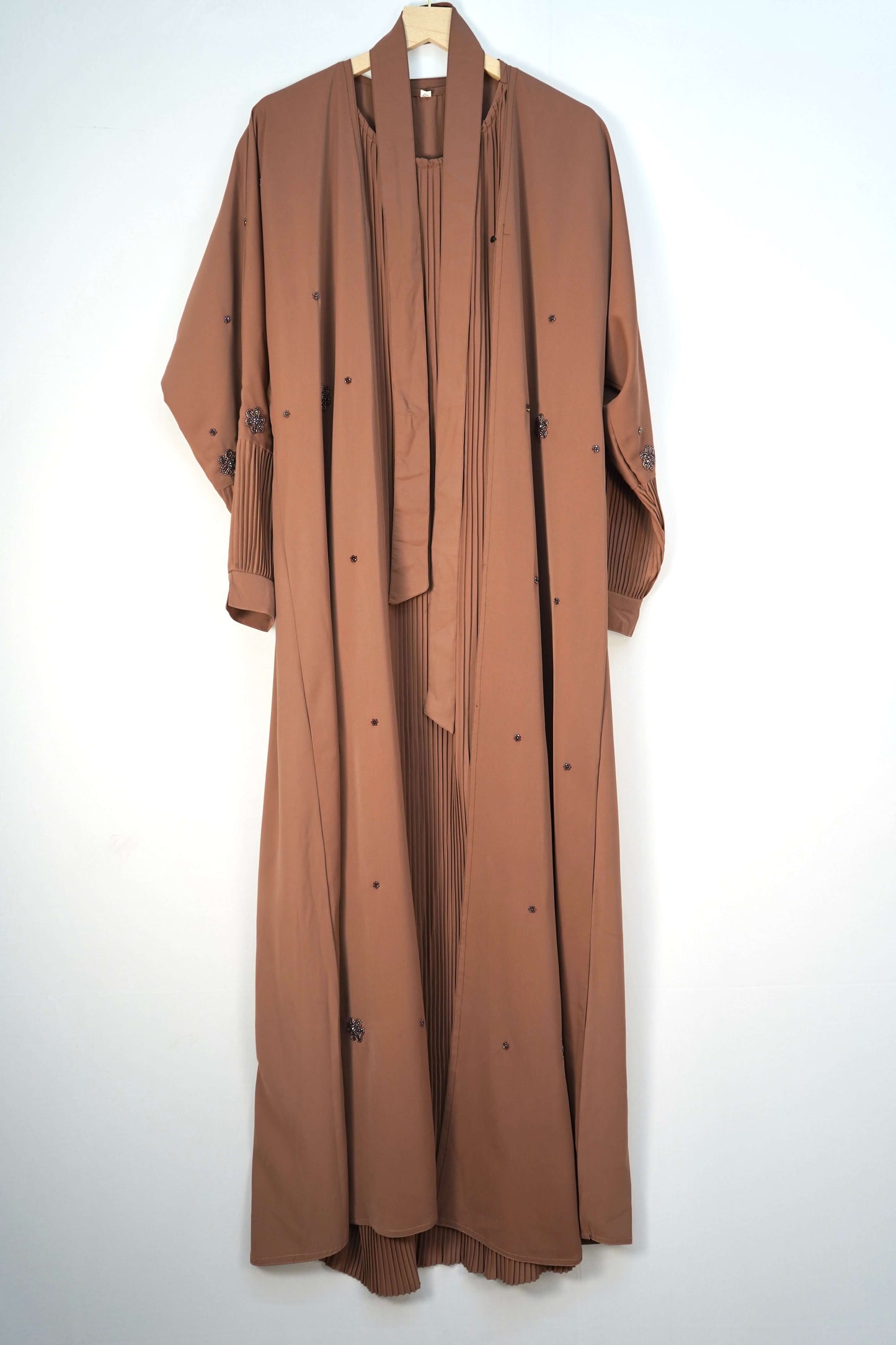 The Korean Alqa Nidha Abaya in brown, hanging open to clearly display the attached belt-like ties alongside the pleated inner layer and beaded details.