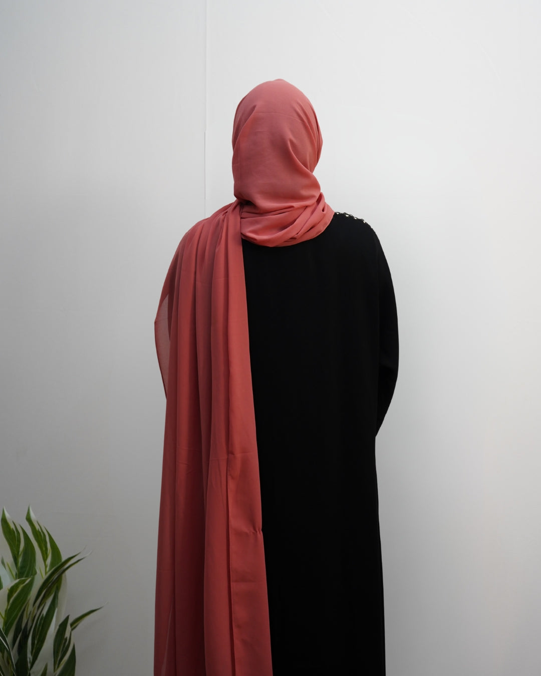 Woman wearing a coral pink chiffon hijab styled with a black abaya, back view.