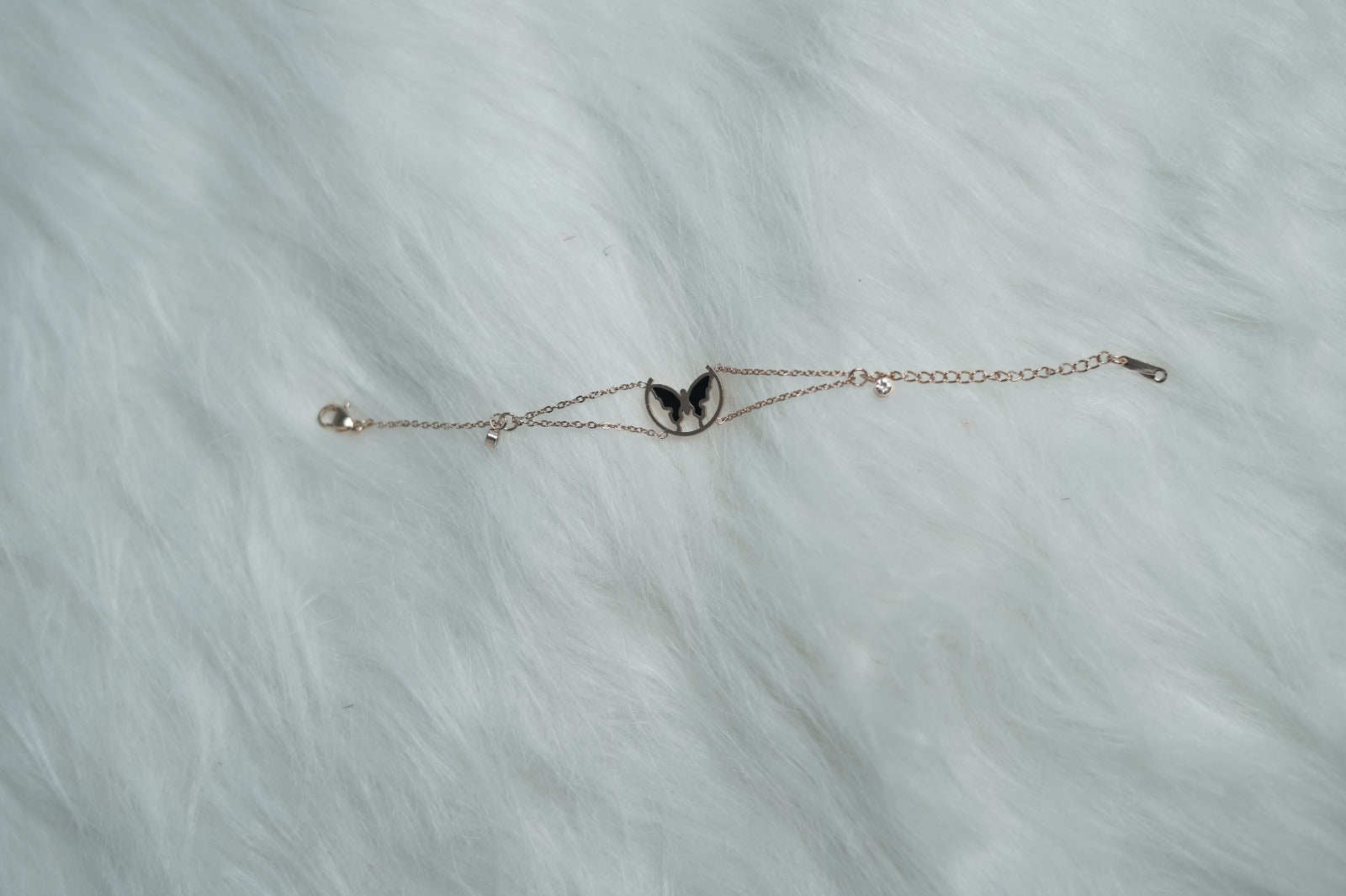 Rose gold-tone bracelet with a butterfly wing charm in a circular frame, laid out on a soft white fur surface.
