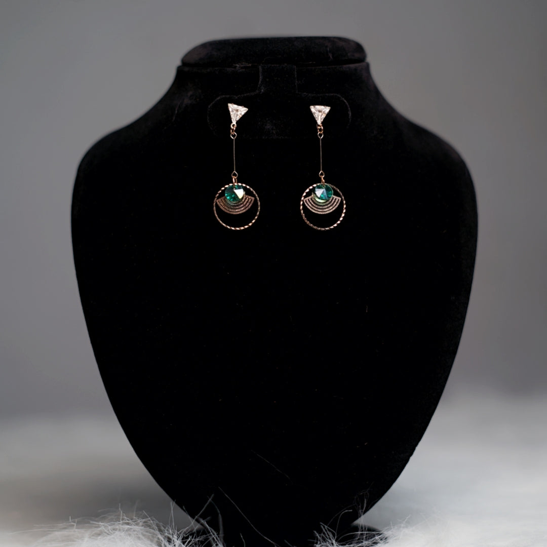 Rose gold geometric drop earrings with emerald green stones and clear triangular studs, displayed on a black velvet stand.