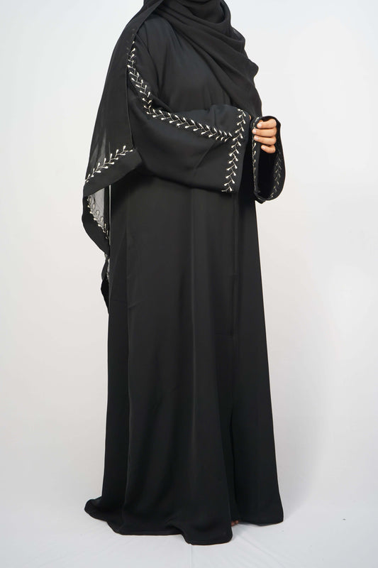 Full shot of a person wearing the black Korean Nidha Abaya with a matching hijab, showing the white leaf-like embroidery on the sleeves and cuff.