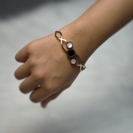 Rose gold bracelet worn on the wrist, featuring a central black circle flanked by two pearl-white accents in a modern swirl layout.