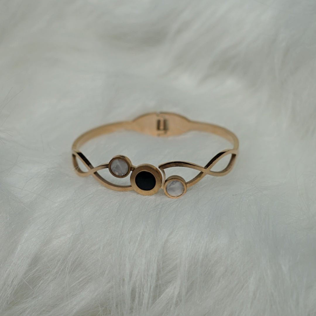 Rose gold bangle with swirl design and three circular stones – black and pearl, displayed on a white fur background.