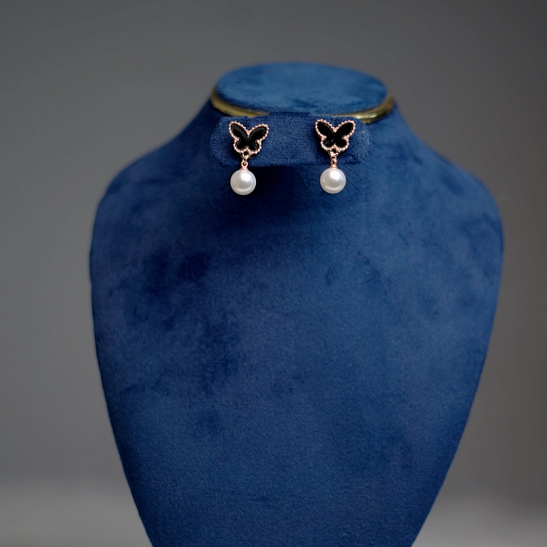 A pair of rose gold-trimmed black butterfly earrings with pearl drops, displayed on a royal blue velvet stand.