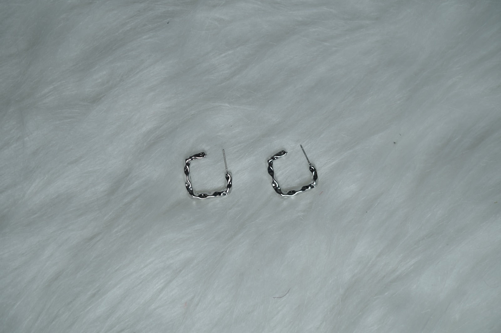 Molten Wave Square Hoops placed on soft white fur background, showcasing wavy silver texture.