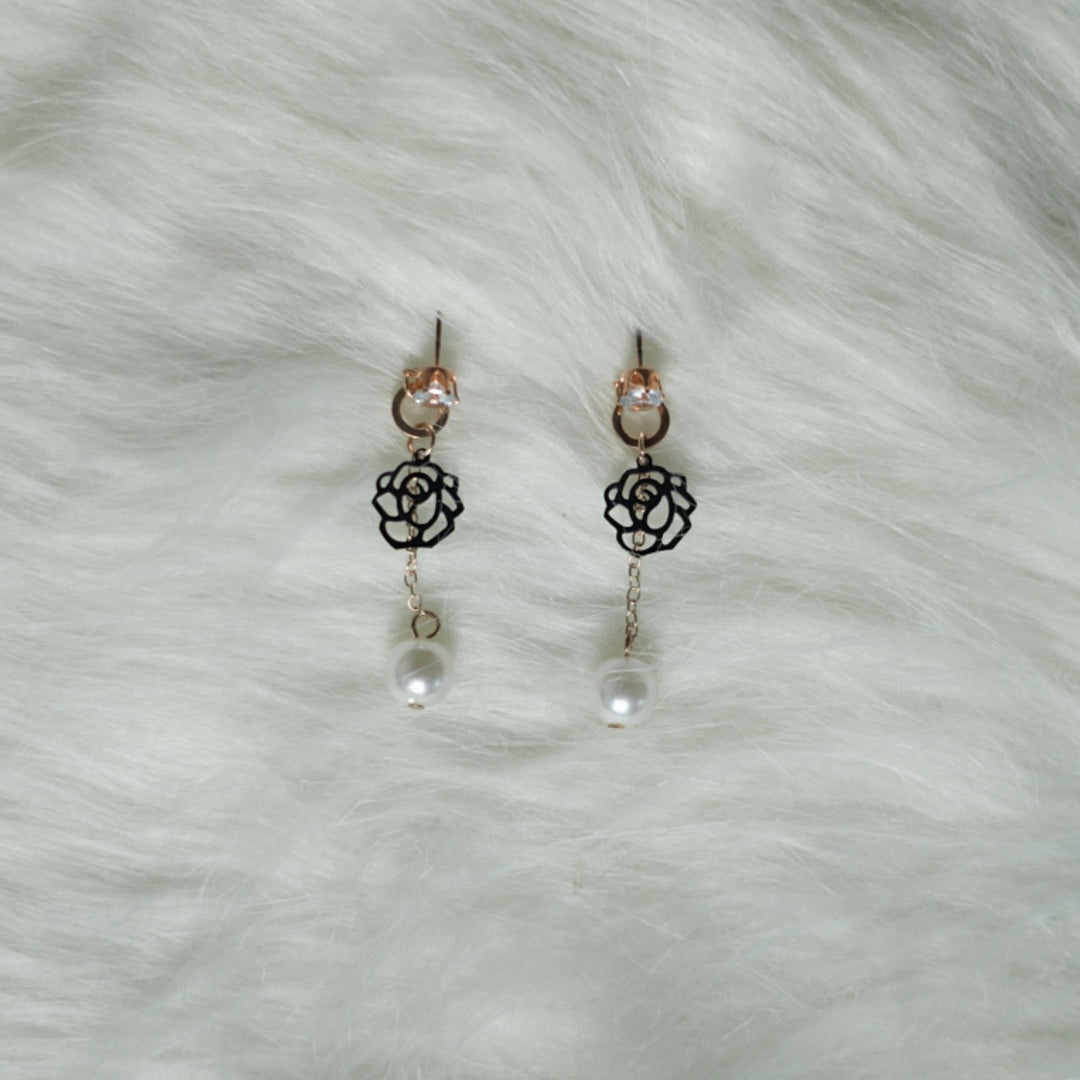 A pair of rose gold earrings with black floral cutouts and pearl drops, placed on a soft white surface.