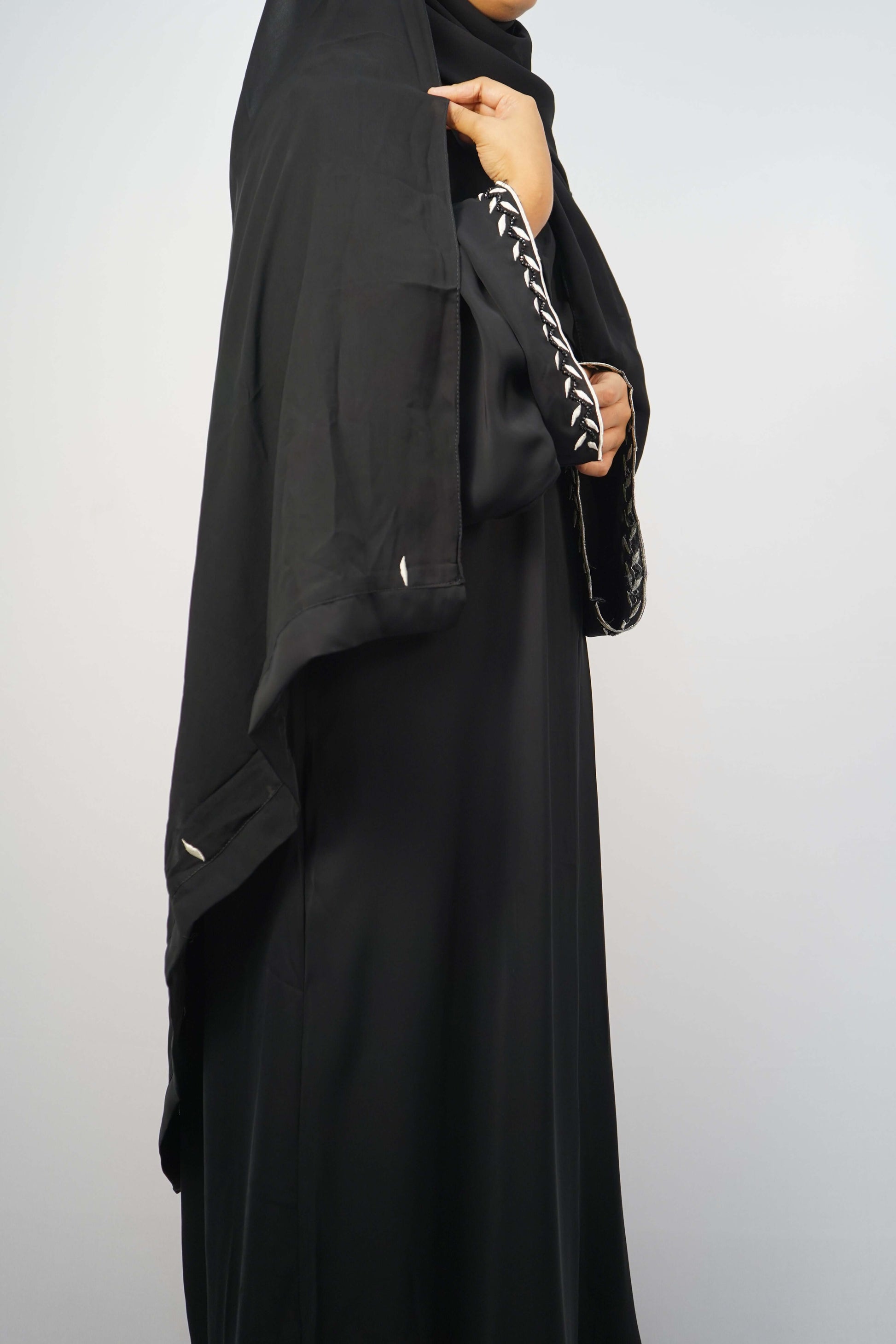 A side view of a woman wearing the black abaya, highlighting the flow of the fabric and the contrasting white embroidery on the hem.