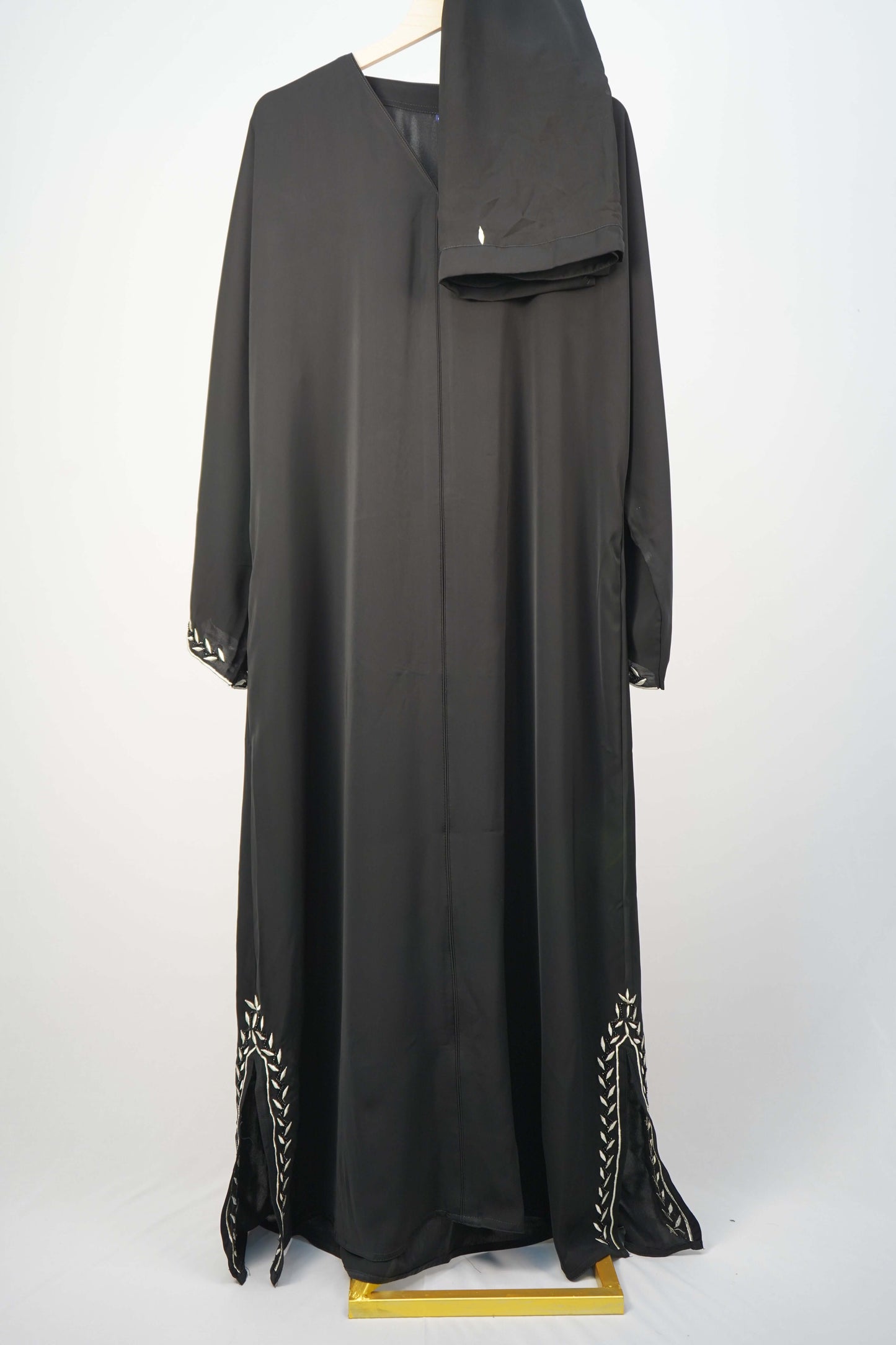 The classic Korean Nidha Abaya in black, hanging with a matching scarf to showcase its full design and contrasting white leaf embroidery.