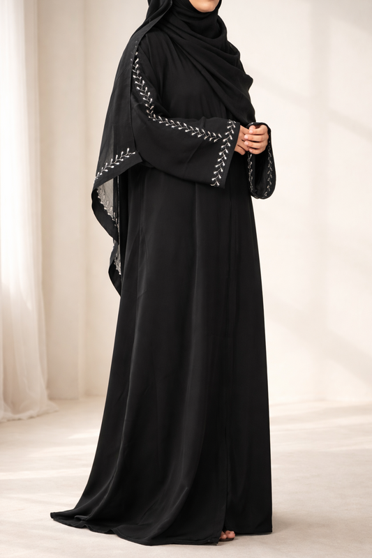 Full shot of a person wearing the black Korean Nidha Abaya with a matching hijab, showing the white leaf-like embroidery on the sleeves and cuff.