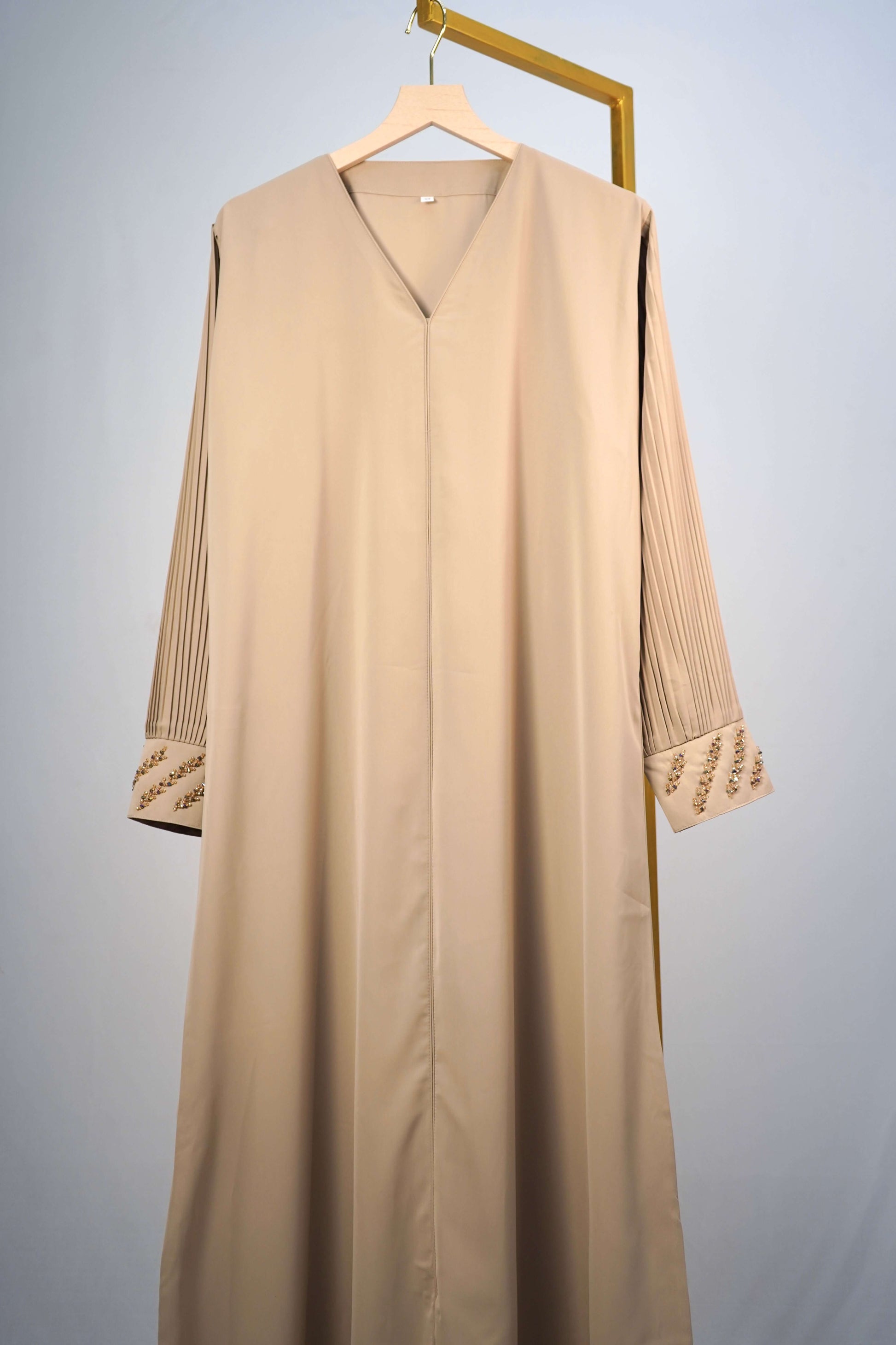 A full shot of the Korean Alqa Nidha Abaya in Light Beige, hanging to highlight its silky fabric, pleated sleeve design and V-neck.