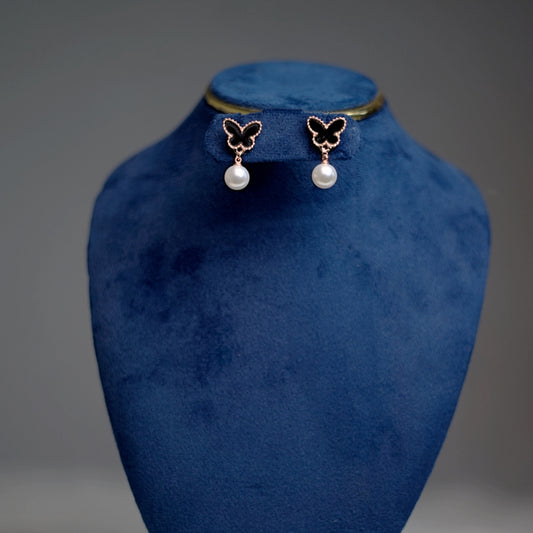 A pair of rose gold-trimmed black butterfly earrings with pearl drops, displayed on a royal blue velvet stand.
